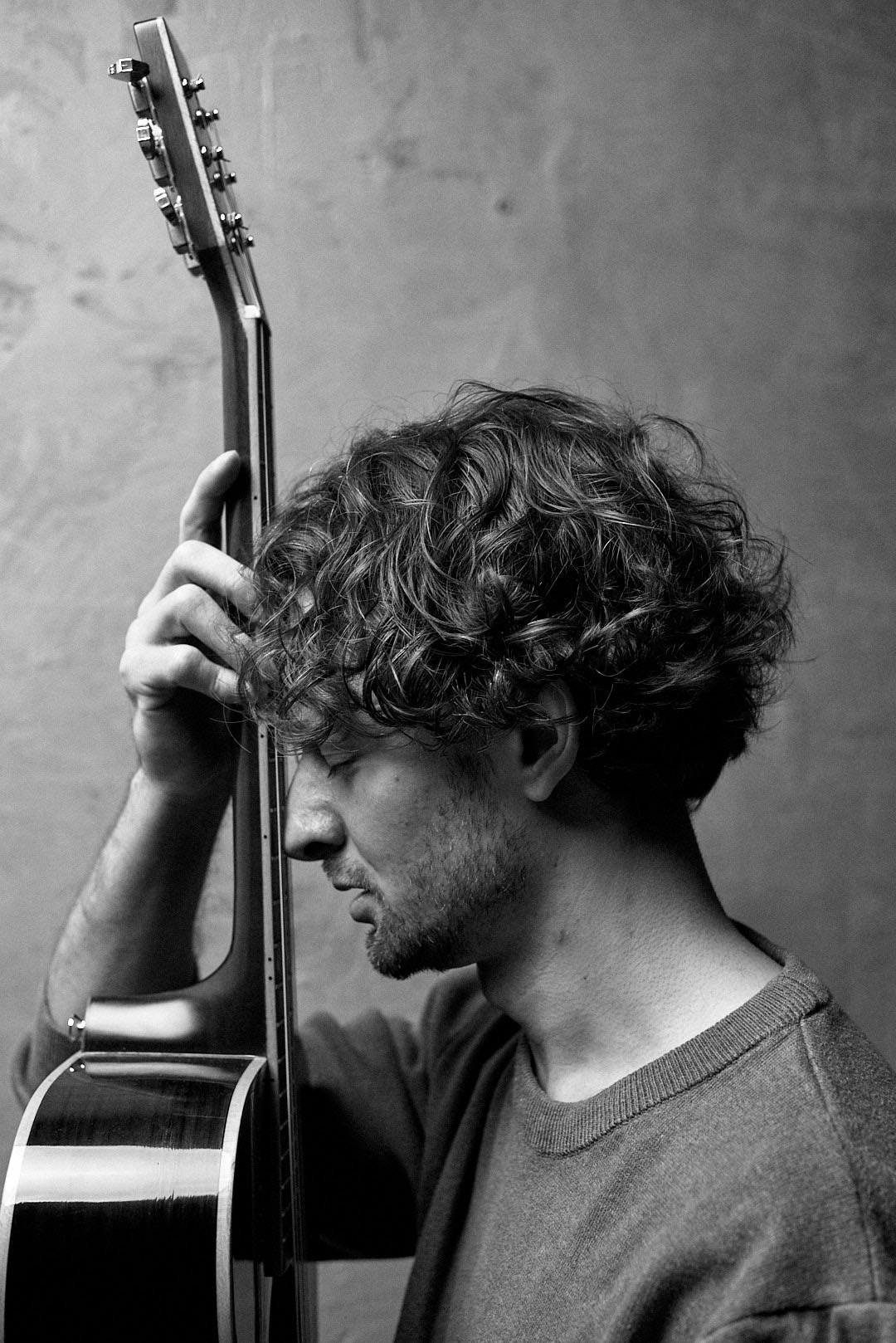 Czarno-biały portret zamyślonego mężczyzny z kręconymi włosami, opierającego głowę o gryf gitary akustycznej, na tle gładkiej ściany.