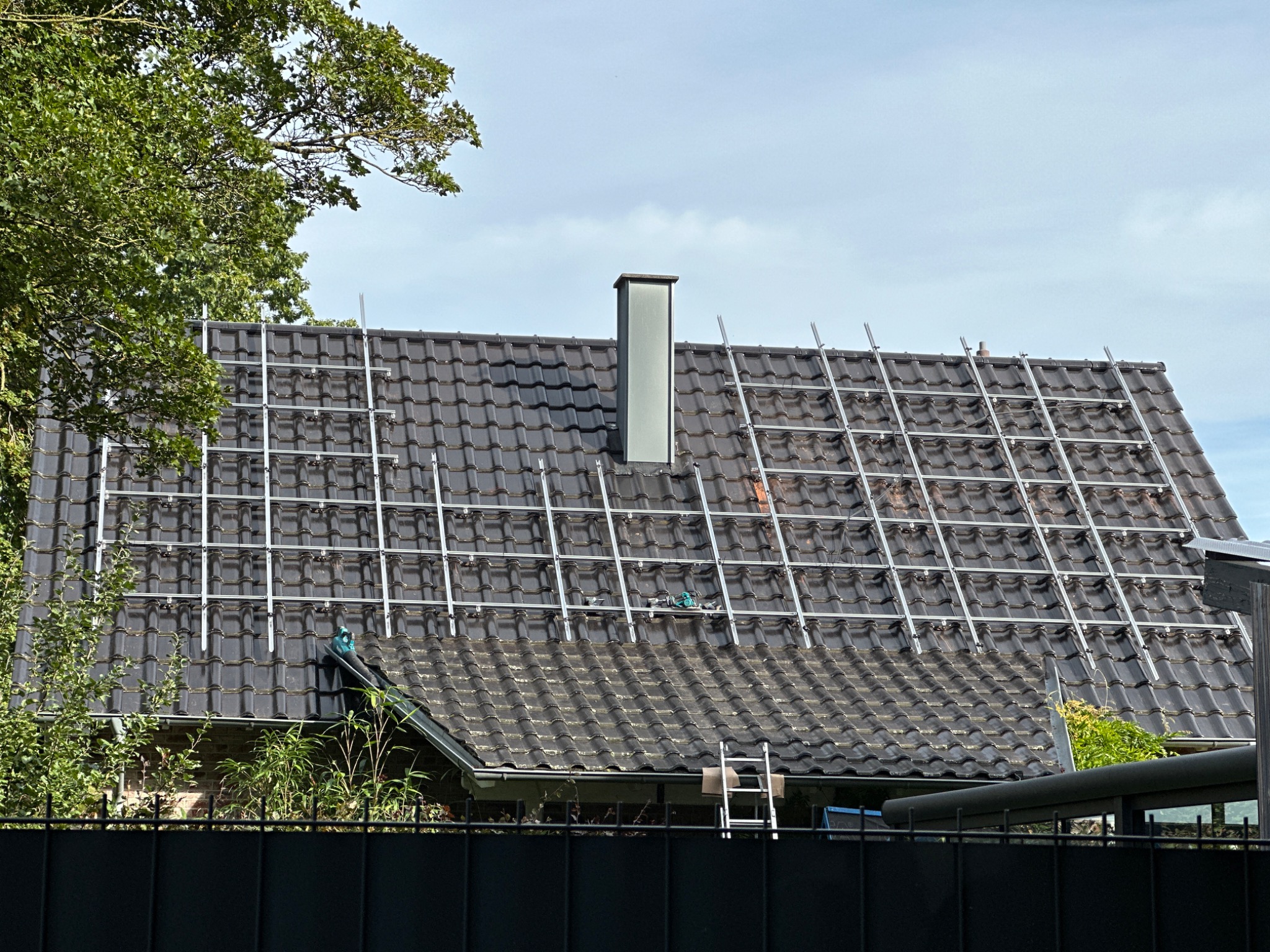 Montaż stelażu pod panele na ciemnym, spadzistym dachu z ceramicznej dachówki, widoczna drabina oparta o rynnę i narzędzia na dachu, komin w tle.