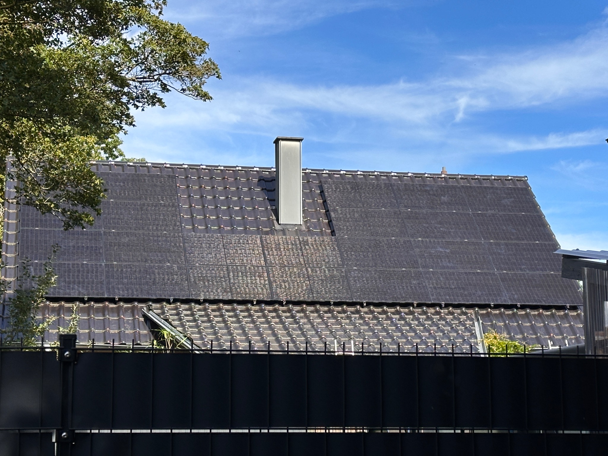 Ciemne panele fotowoltaiczne zintegrowane z dachówką na dachu domu jednorodzinnego, widoczne zza czarnego ogrodzenia i drzewa, z kominem w tle, błękitne niebo.