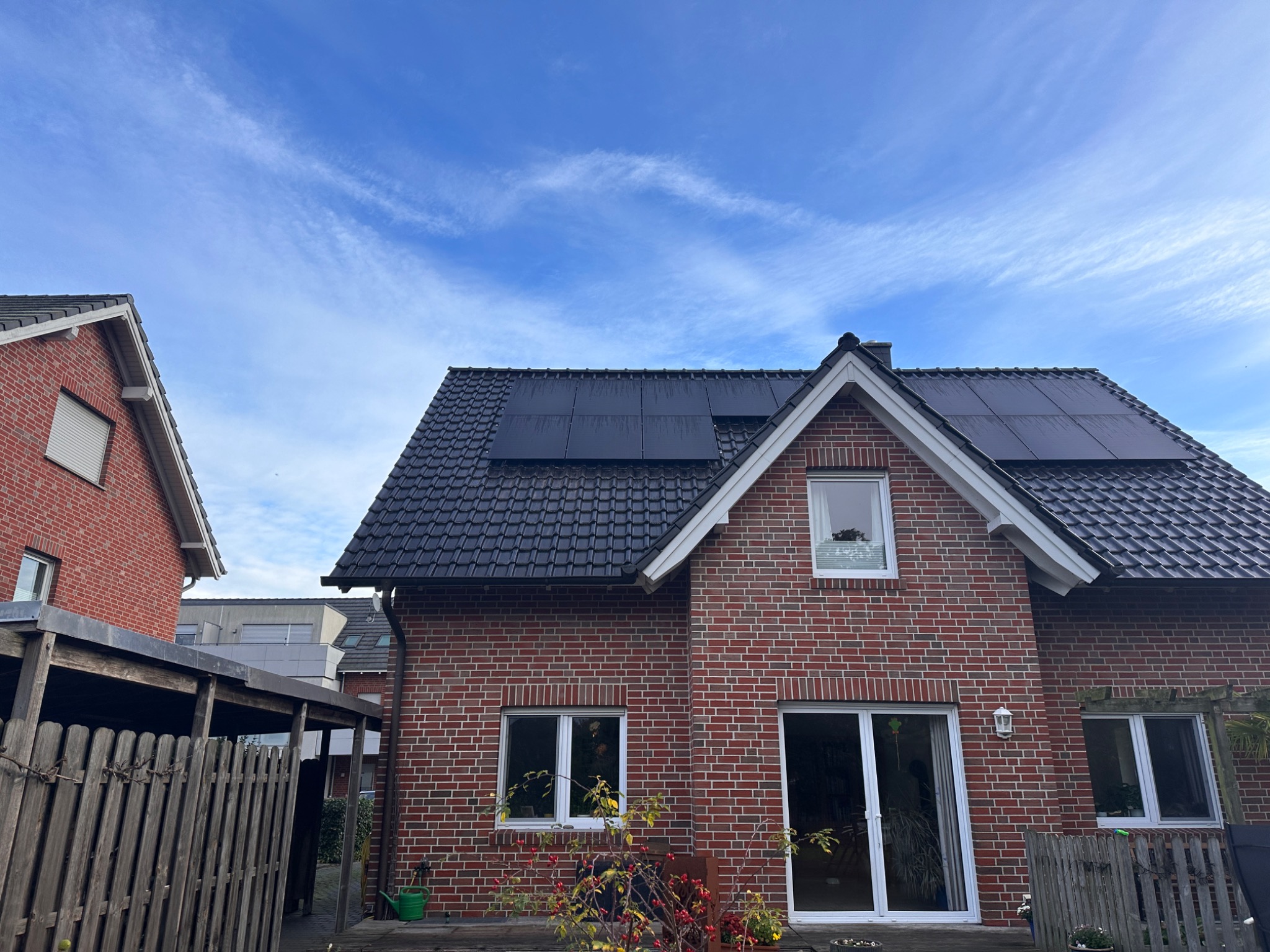 Dom jednorodzinny z ciemnymi panelami słonecznymi na dachu, widok z dołu na tle błękitnego nieba z chmurami.