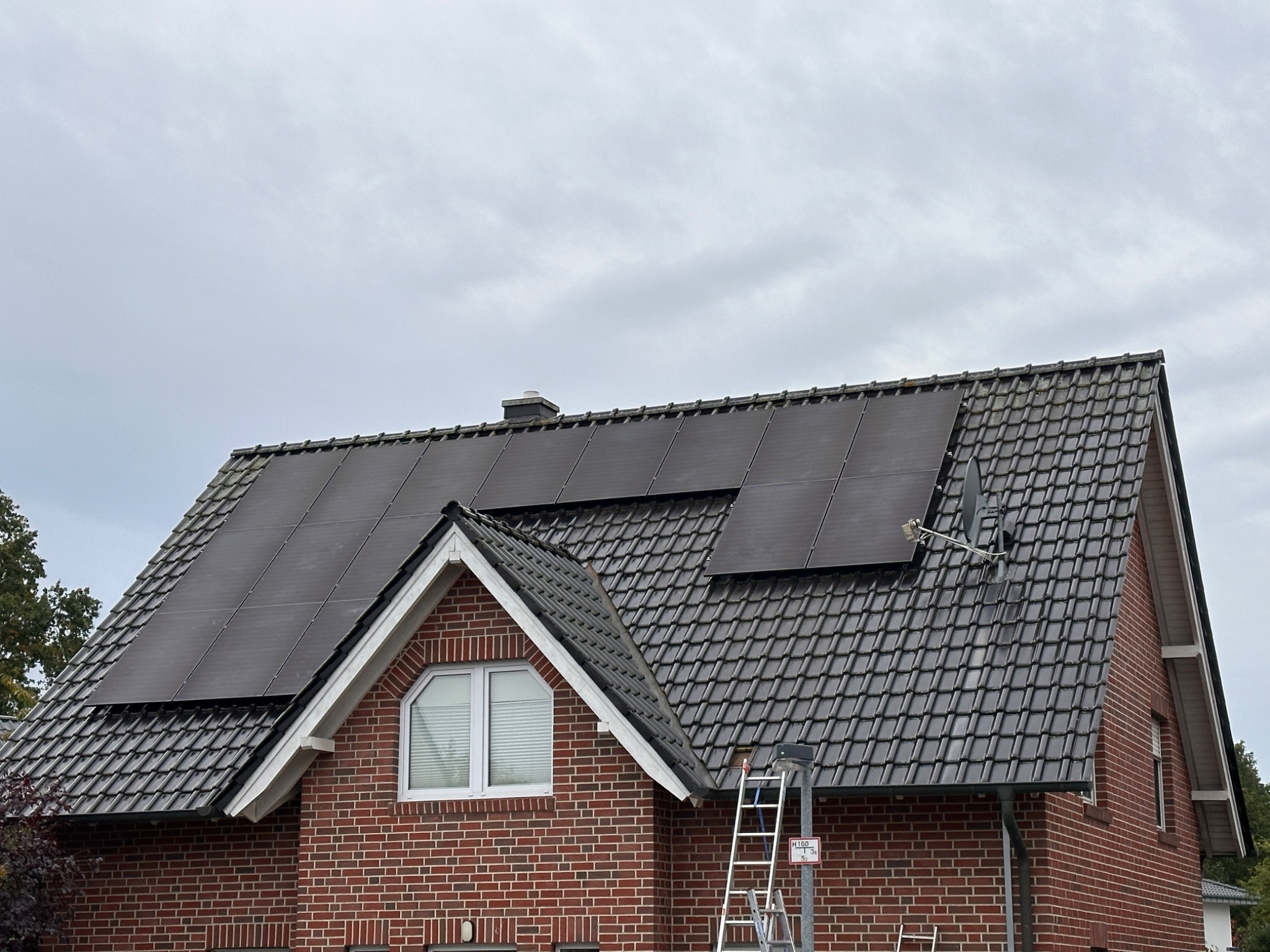 Częściowo zainstalowane panele słoneczne na dachu domu z czerwonej cegły, drabina oparta o dach, pochmurne niebo.