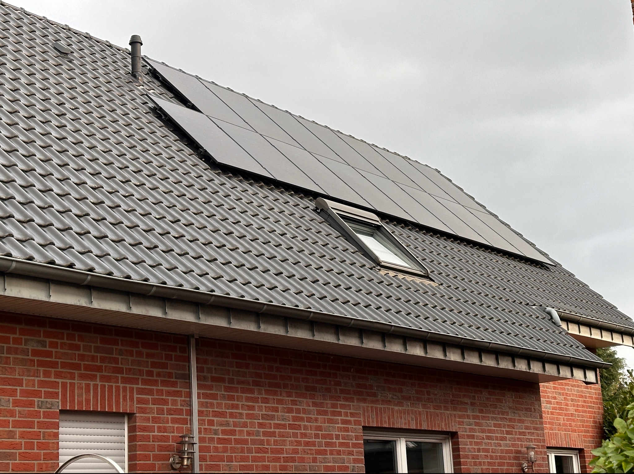 Ciemne panele słoneczne na szarym, spadzistym dachu z dachówki ceramicznej, obok okna dachowego i komina wentylacyjnego, na tle pochmurnego nieba.