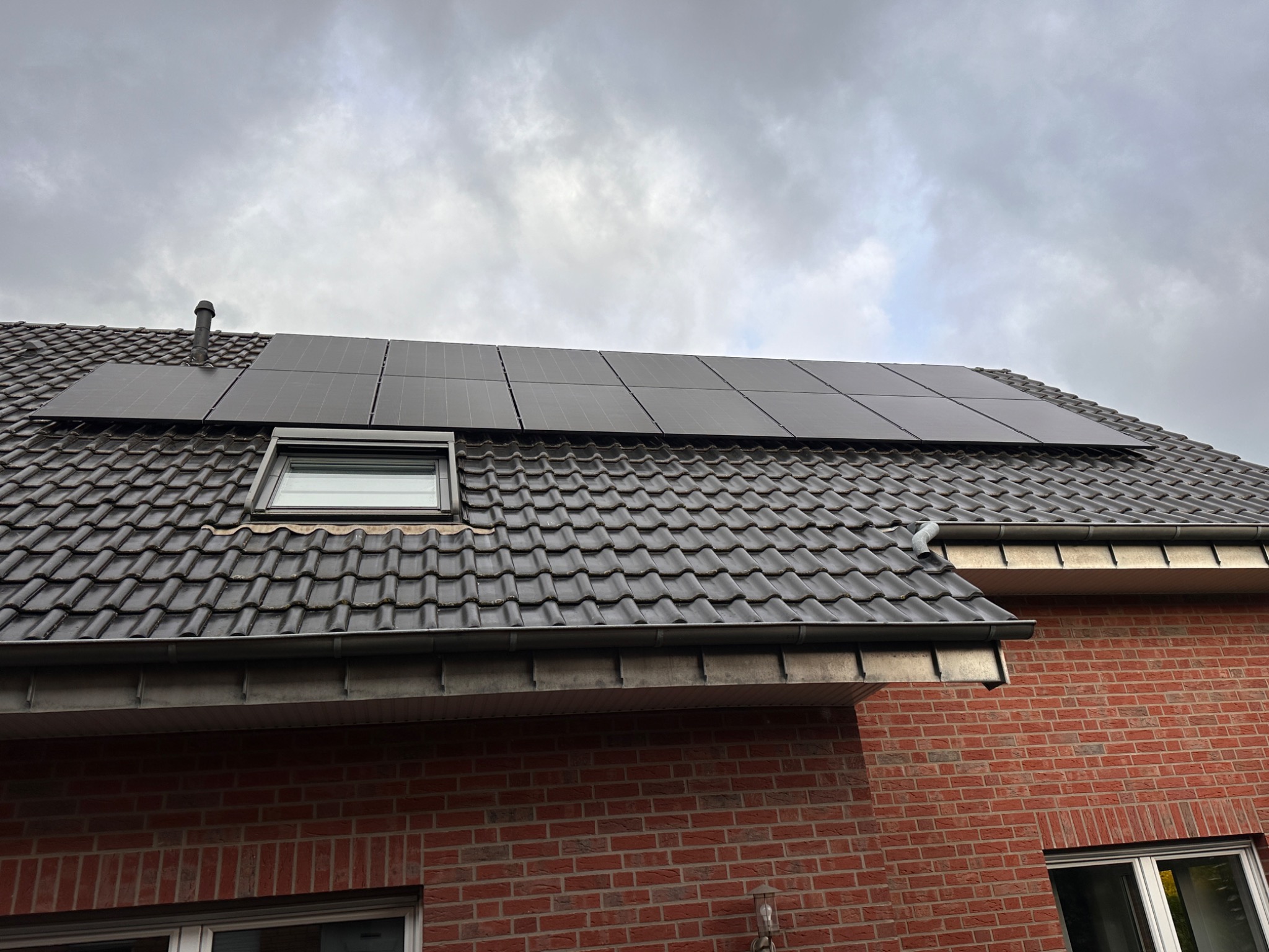 Czarny panel fotowoltaiczny na grafitowym dachu z dachówki ceramicznej, widoczny komin wentylacyjny i okno dachowe, fragment ściany z czerwonej cegły