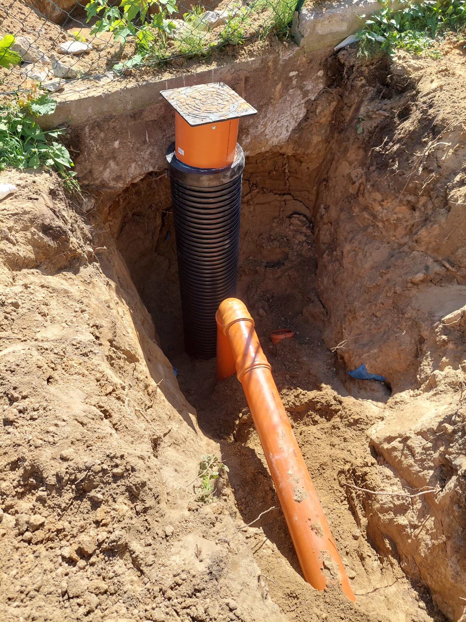 Wykop w ziemi z widoczną czarną rurą karbowaną pionowo, pomarańczową rurą pod kątem i pomarańczową studzienką rewizyjną z pokrywą na górze, przy ogrodzeniu z siatki.