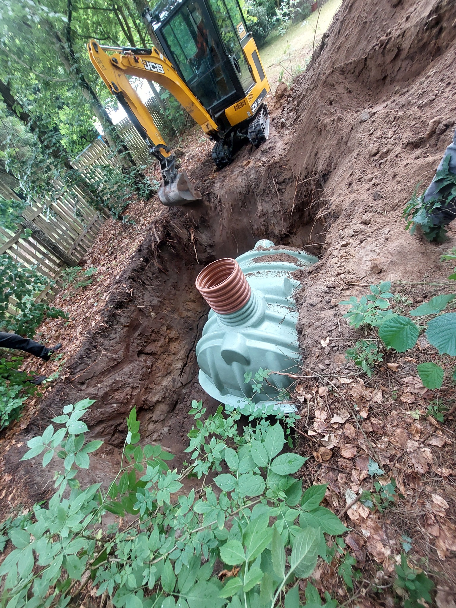 Żółta koparka JCB umieszcza zielony, plastikowy zbiornik z karbowanym wlotem w wykopie ziemnym na zalesionej działce, widoczne liście i drewniane ogrodzenie.