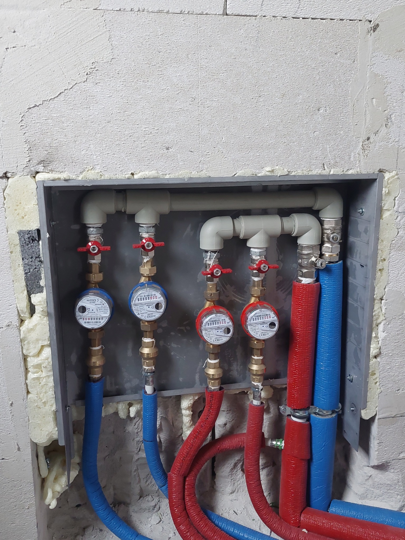 Wnęka z czterema wodomierzami, rurami w izolacji czerwonej i niebieskiej, oraz zaworami z czerwonymi pokrętłami, zamontowana w surowej ścianie z widoczną pianką montażową.