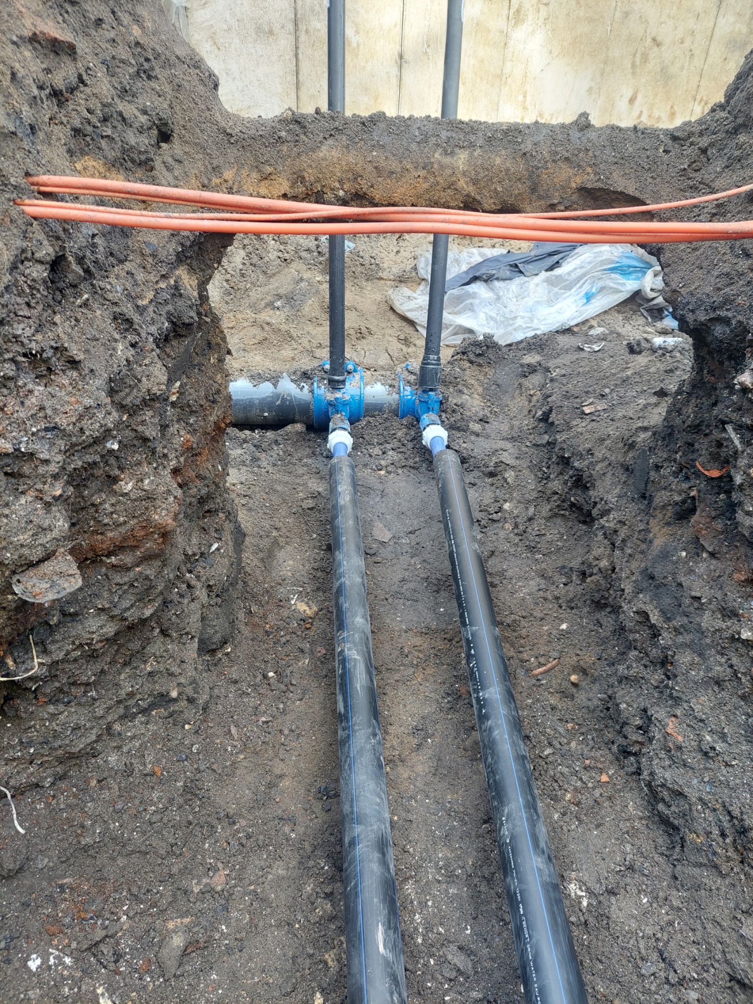 Dwie czarne rury wodociągowe z niebieskim oznaczeniem, połączone niebieskim zaworem w wykopie ziemnym, z widocznymi pomarańczowymi kablami w górnej części wykopu.
