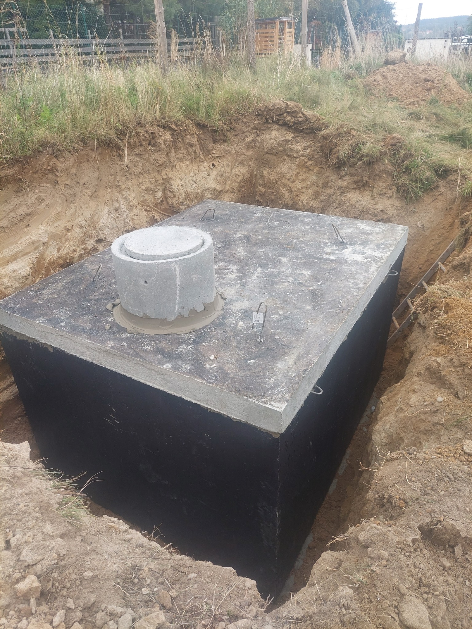 Szambo betonowe, prostokątne, z betonową pokrywą i kominkiem, umieszczone w wykopie ziemnym, zabezpieczone czarną izolacją.