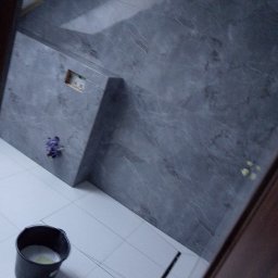 Adam Rączka - Fragment ściany w łazience wyłożonej szarą płytką imitującą marmur, z widocznym odpływem liniowym w podłodze z białych płytek i wiaderkiem z resztkami zaprawy.