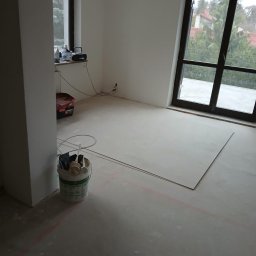 Adam Rączka - Wnętrze w trakcie remontu: surowy beton, płyty karton-gips, wiadro z narzędziami, okno z widokiem na zieleń. Przygotowanie do wykończenia podłogi.