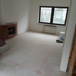 Adam Rączka - Pokój w trakcie prac przygotowawczych: zagruntowana podłoga z wyznaczonymi liniami, widoczny kominek, okno z kratami i grzejnik. Przygotowania do dalszych etapów.