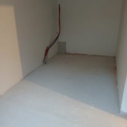 Adam Rączka - Surowe wnętrze pokoju po szpachlowaniu ścian i wylewce podłogi, z widocznymi przewodami elektrycznymi przy ścianie. Przygotowanie do dalszych prac wykończeniowych.