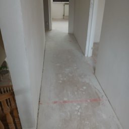 Adam Rączka - Korytarz w trakcie prac wykończeniowych. Widoczne ściany po szpachlowaniu i podłoga z resztkami zaprawy. W tle okno i fragment schodów z drewnianą balustradą.