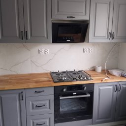 Adam Rączka - Szare szafki kuchenne z drewnianym blatem i czarnym piekarnikiem. Marmurowe płytki na ścianie z gniazdkami. Ukończona zabudowa kuchenna.