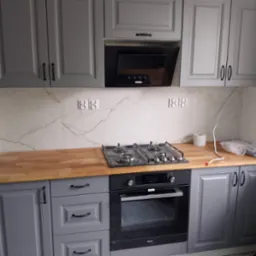 Szare szafki kuchenne z drewnianym blatem i czarnym piekarnikiem. Marmurowe płytki na ścianie z gniazdkami. Ukończona zabudowa kuchenna.