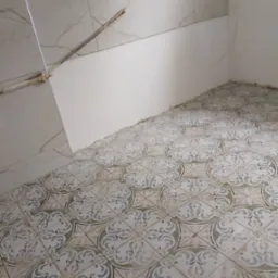 Ułożone płytki podłogowe z dekoracyjnym wzorem w łazience, częściowo wykończona ściana z marmurowymi płytkami i instalacją elektryczną. Widok z góry.