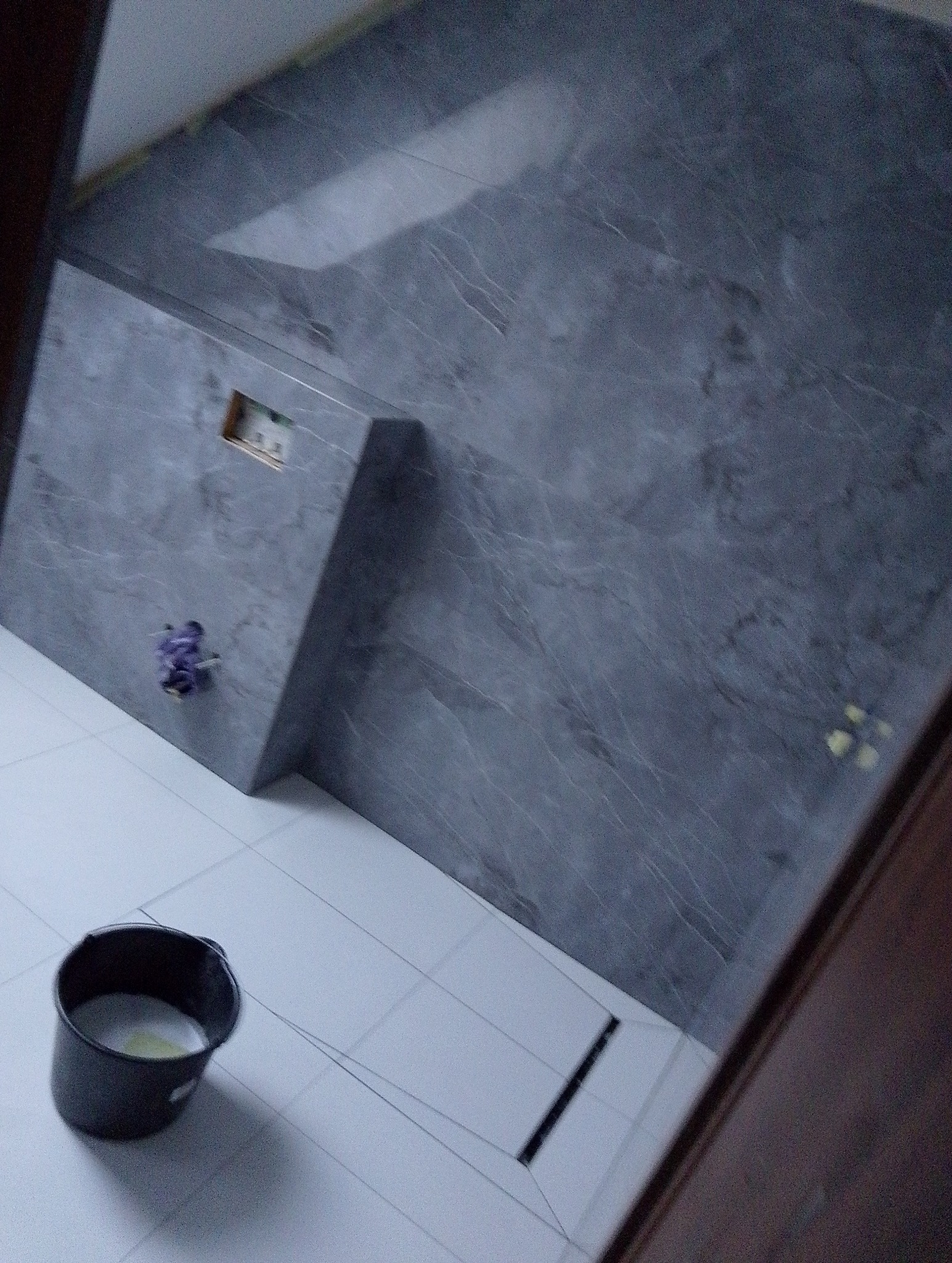 Fragment ściany w łazience wyłożonej szarą płytką imitującą marmur, z widocznym odpływem liniowym w podłodze z białych płytek i wiaderkiem z resztkami zaprawy.