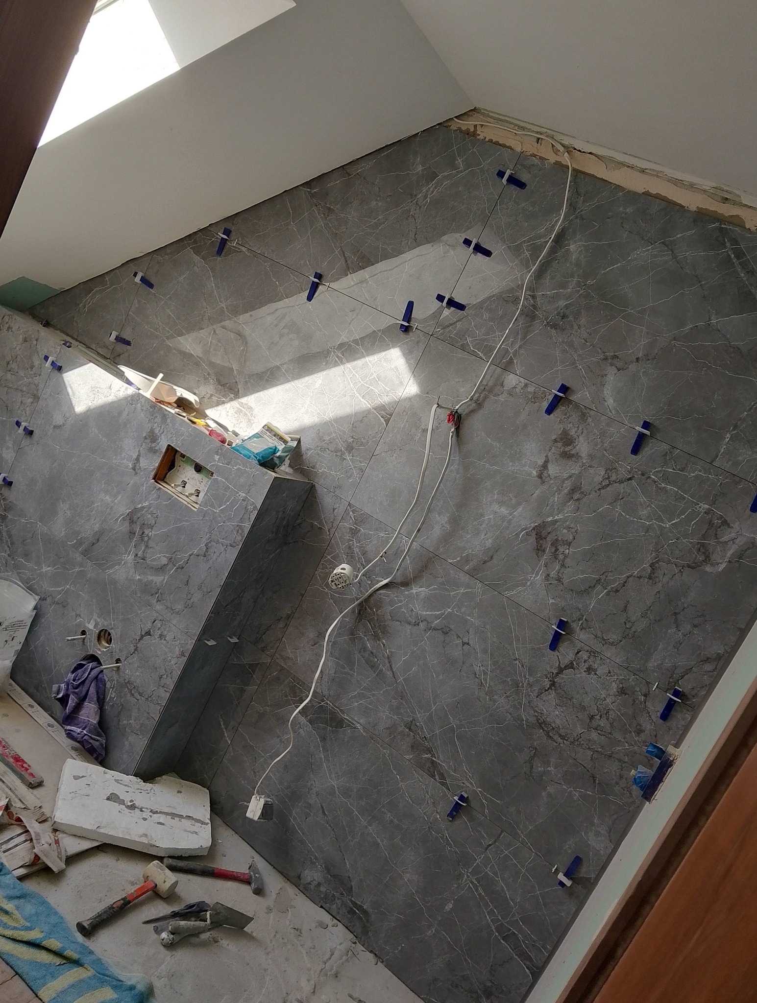 Świeżo ułożone, marmurowe płytki na ścianie łazienki, zabezpieczone niebieskimi klipsami poziomującymi, narzędzia glazurnicze i resztki zaprawy na podłodze, widok z góry.