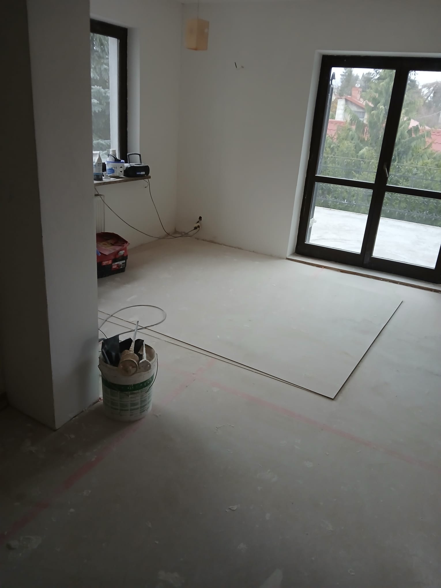 Wnętrze w trakcie remontu: surowy beton, płyty karton-gips, wiadro z narzędziami, okno z widokiem na zieleń. Przygotowanie do wykończenia podłogi.