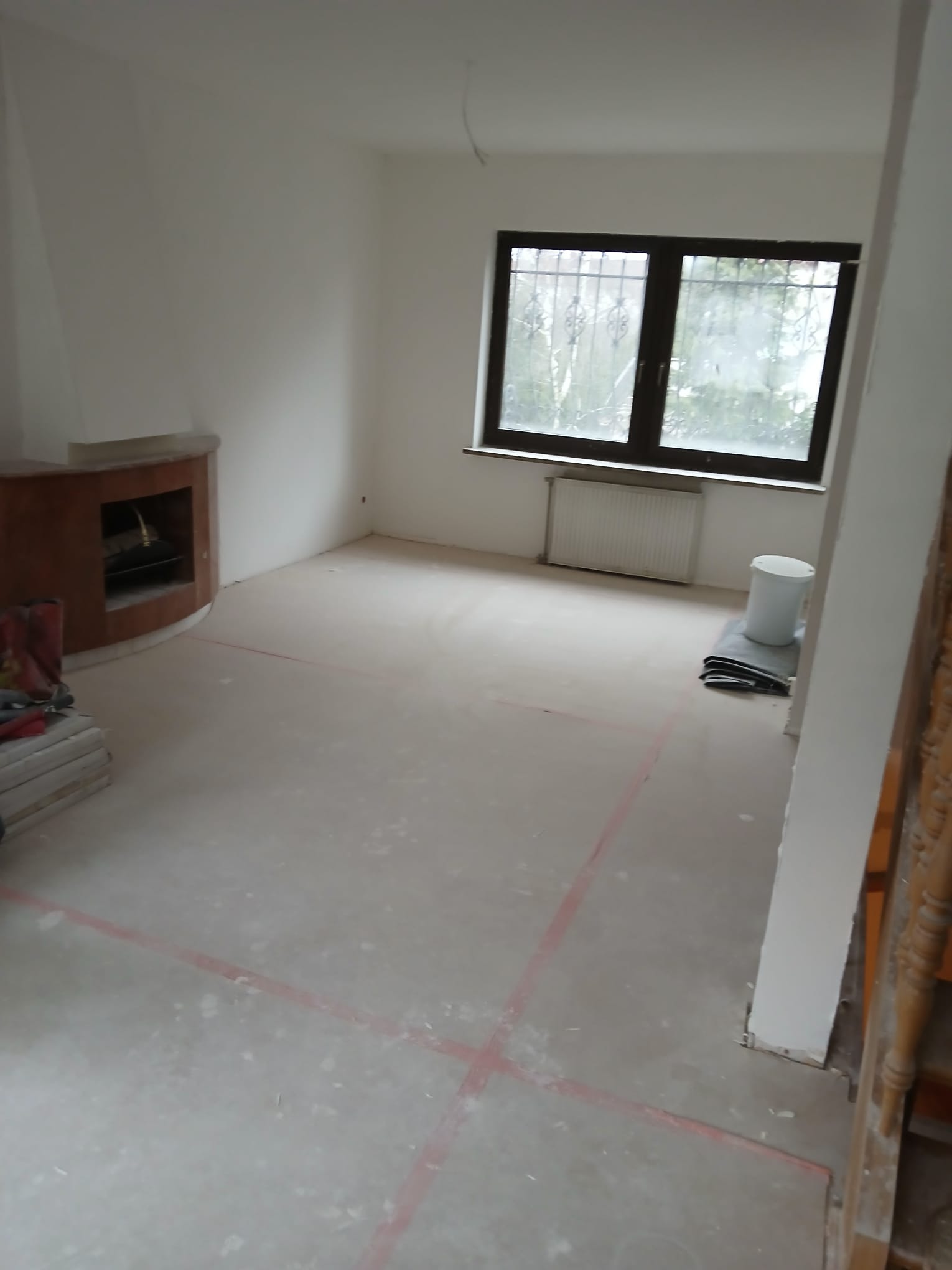 Pokój w trakcie prac przygotowawczych: zagruntowana podłoga z wyznaczonymi liniami, widoczny kominek, okno z kratami i grzejnik. Przygotowania do dalszych etapów.