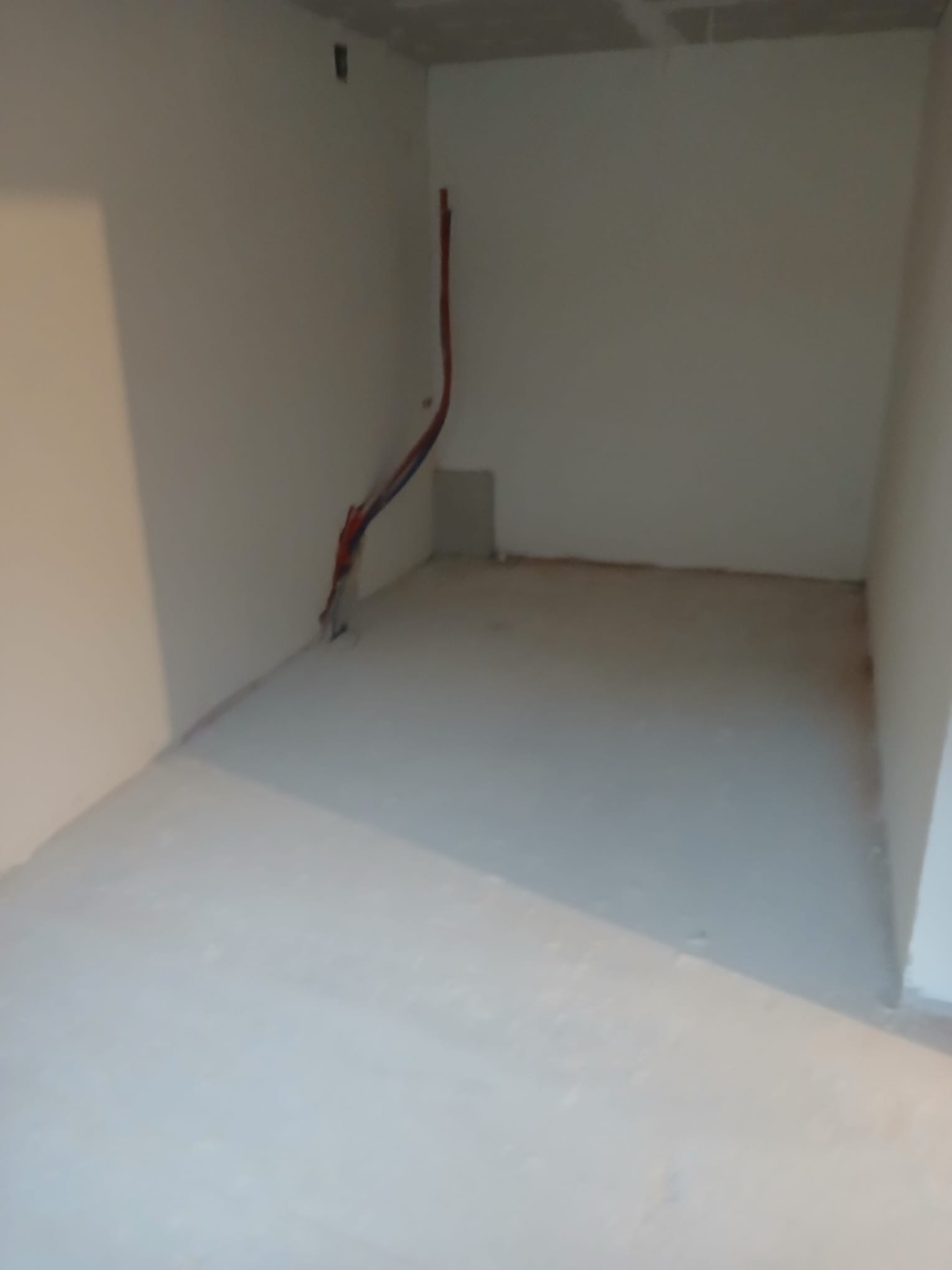 Surowe wnętrze pokoju po szpachlowaniu ścian i wylewce podłogi, z widocznymi przewodami elektrycznymi przy ścianie. Przygotowanie do dalszych prac wykończeniowych.