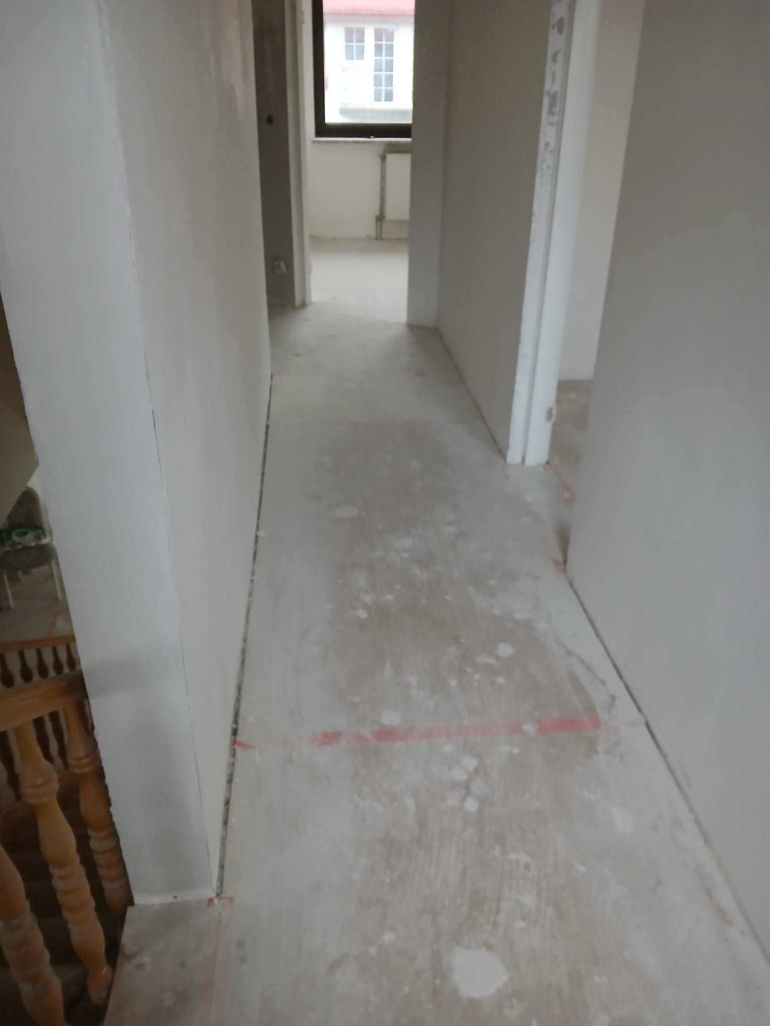 Korytarz w trakcie prac wykończeniowych. Widoczne ściany po szpachlowaniu i podłoga z resztkami zaprawy. W tle okno i fragment schodów z drewnianą balustradą.