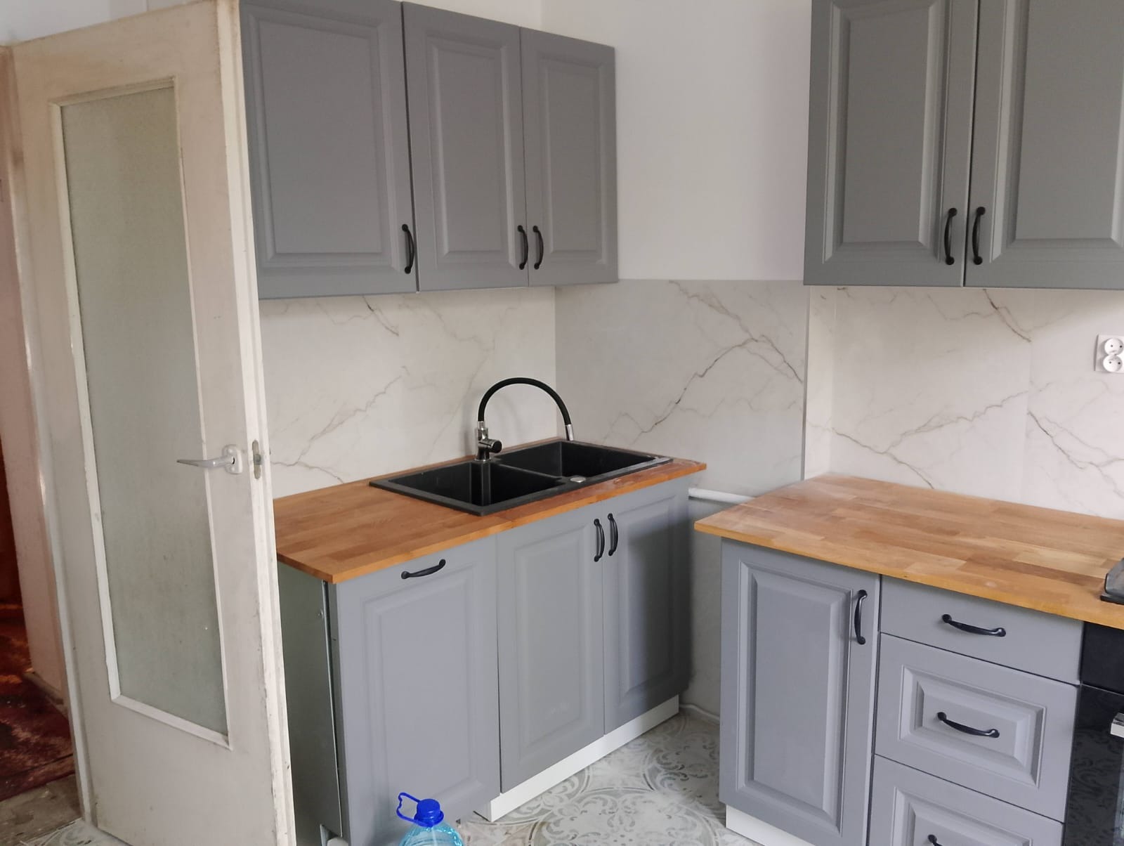 Szare szafki kuchenne z drewnianym blatem i marmurowymi płytkami, widok z otwartymi drzwiami. Ujęcie prezentuje detale wykończenia.