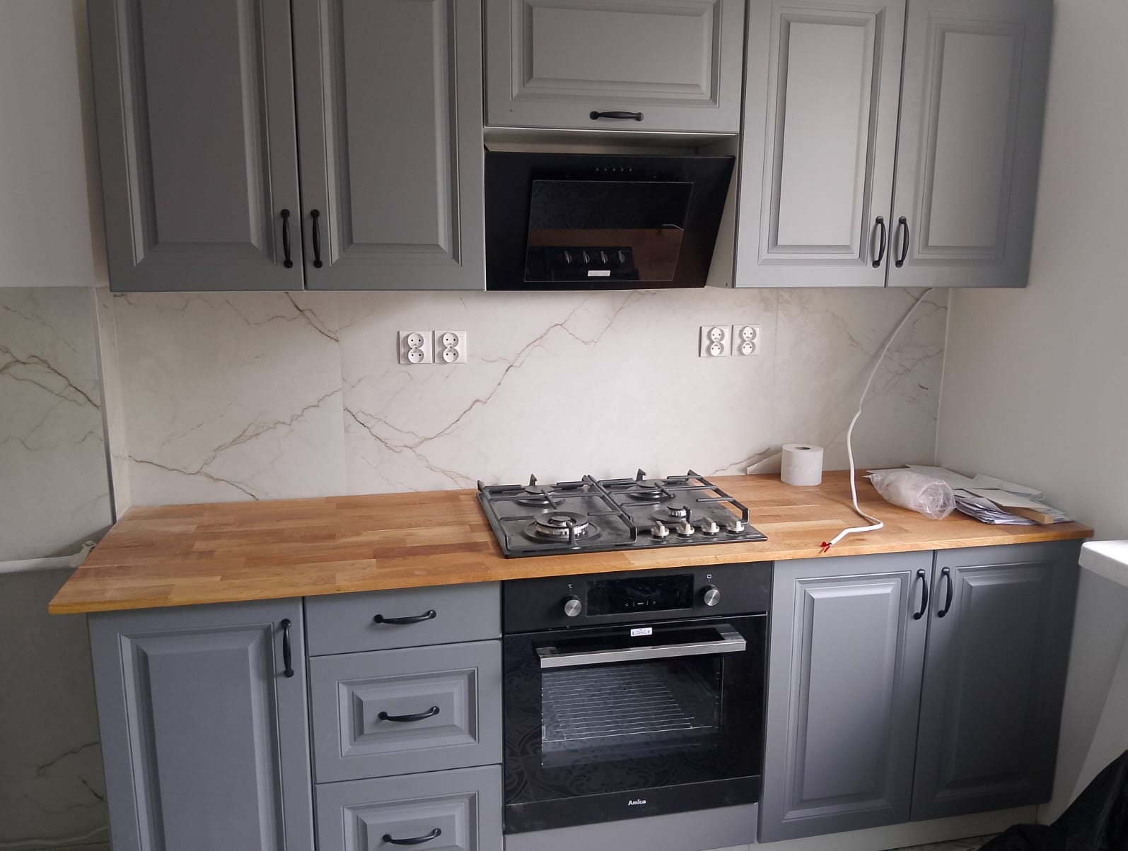 Szare szafki kuchenne z drewnianym blatem i czarnym piekarnikiem. Marmurowe płytki na ścianie z gniazdkami. Ukończona zabudowa kuchenna.