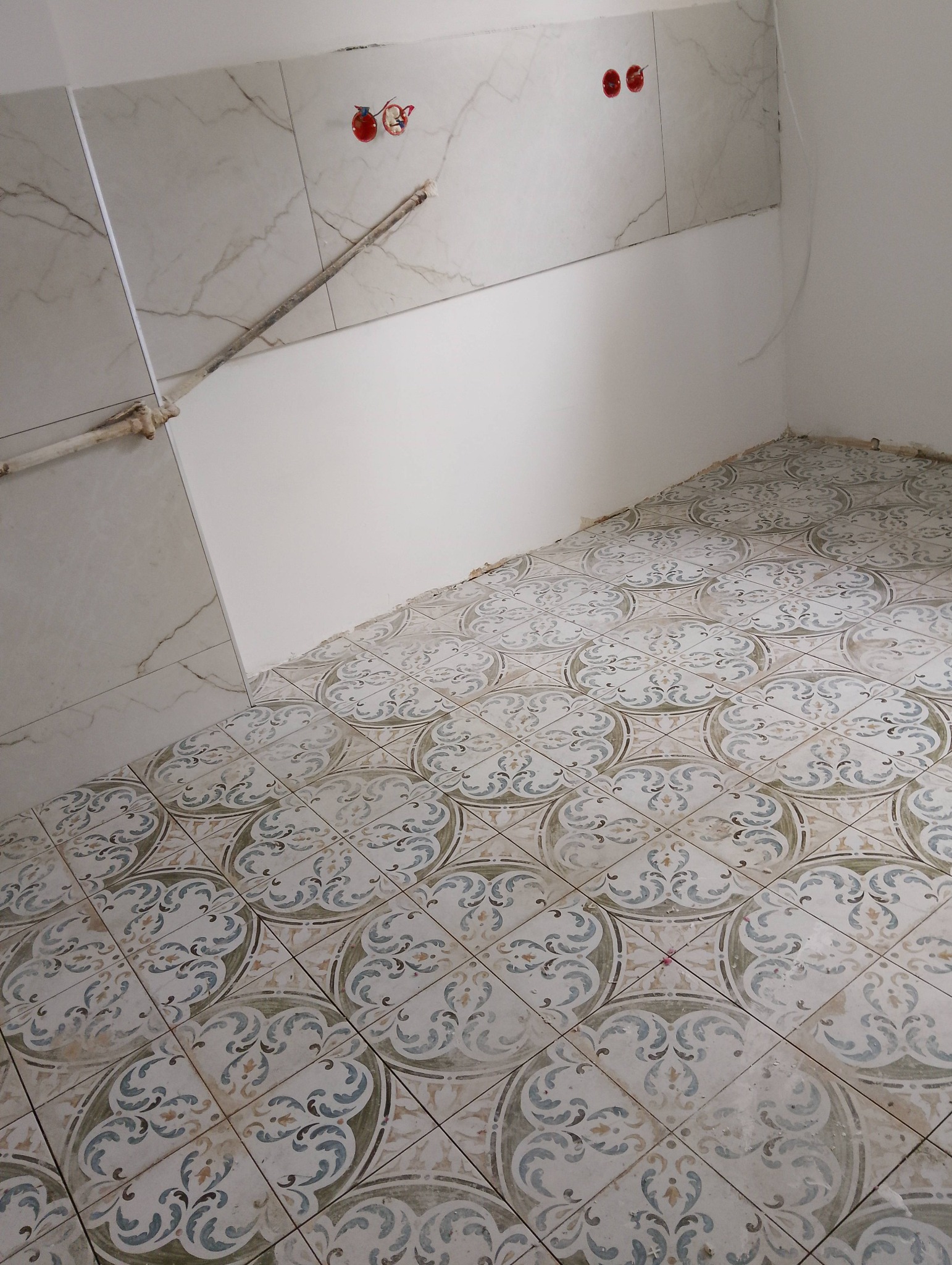 Ułożone płytki podłogowe z dekoracyjnym wzorem w łazience, częściowo wykończona ściana z marmurowymi płytkami i instalacją elektryczną. Widok z góry.