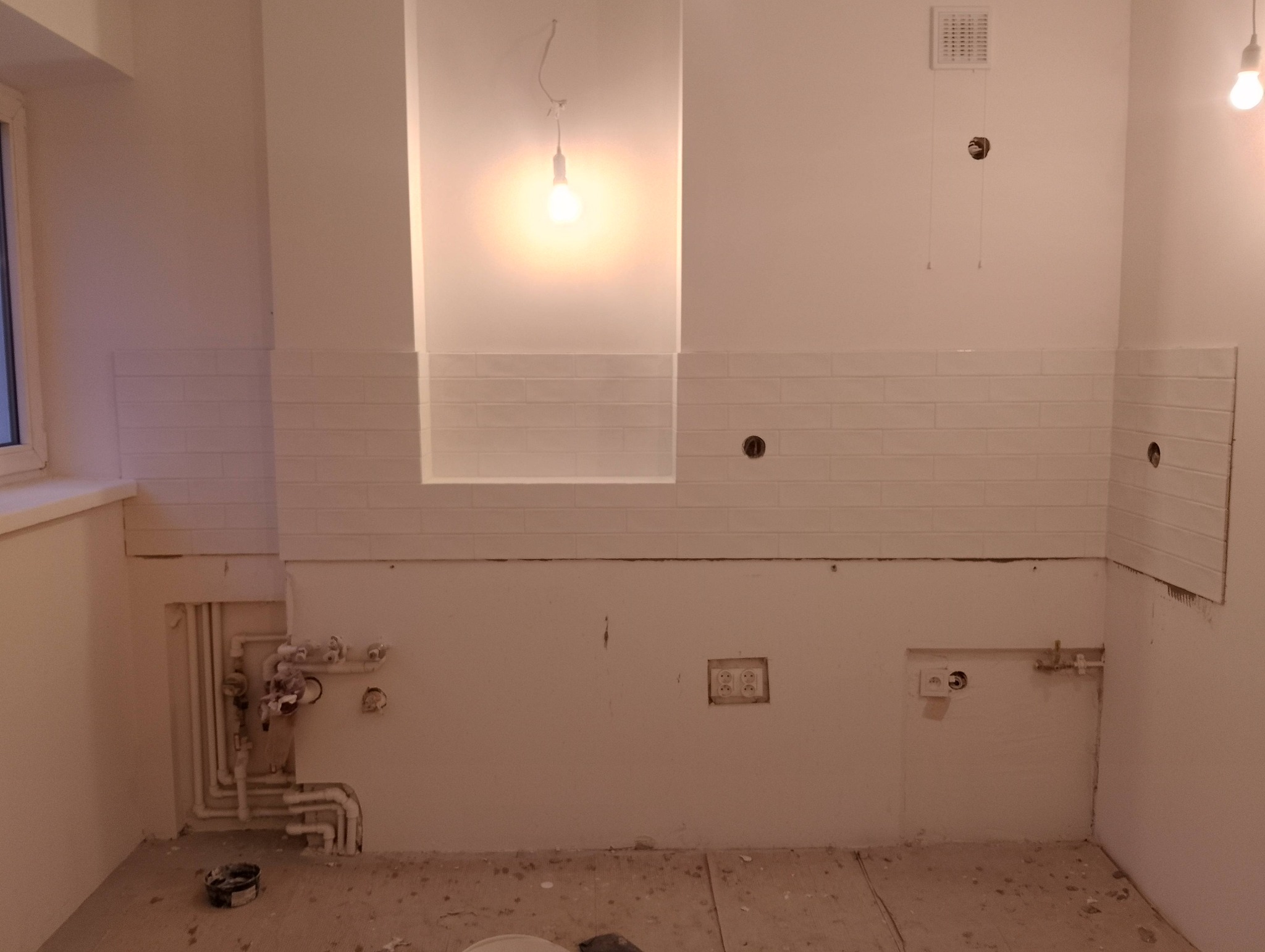 Biała kuchnia w trakcie remontu: częściowo ułożone białe płytki typu metro, widoczne instalacje, gniazdka elektryczne i surowy stan podłogi. Oświetlenie punktowe.