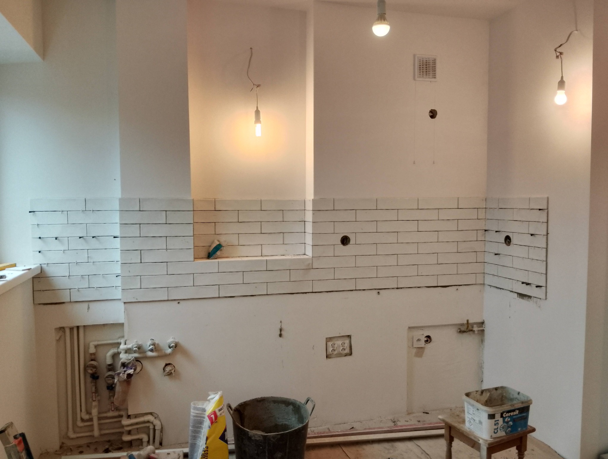 Białe, prostokątne płytki częściowo ułożone na ścianie w trakcie remontu. Widoczne narzędzia, wiadro i worki z zaprawą. Surowe oświetlenie żarówkami.