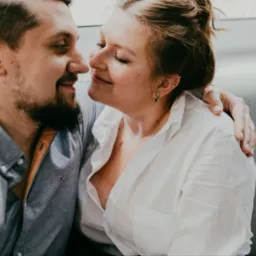 Uśmiechnięta para, on z brodą w szarej koszuli, ona w białej, obejmująca się czule, zbliżająca się do pocałunku, z kolczykiem typu helix w uchu, na neutralnym tle.