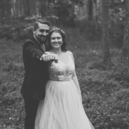 Czarno-białe zdjęcie uśmiechniętej pary młodej w lesie; Panna Młoda w sukni z koronkową górą i wiankiem, Pan Młody w garniturze, oboje wskazują w stronę obiektywu.