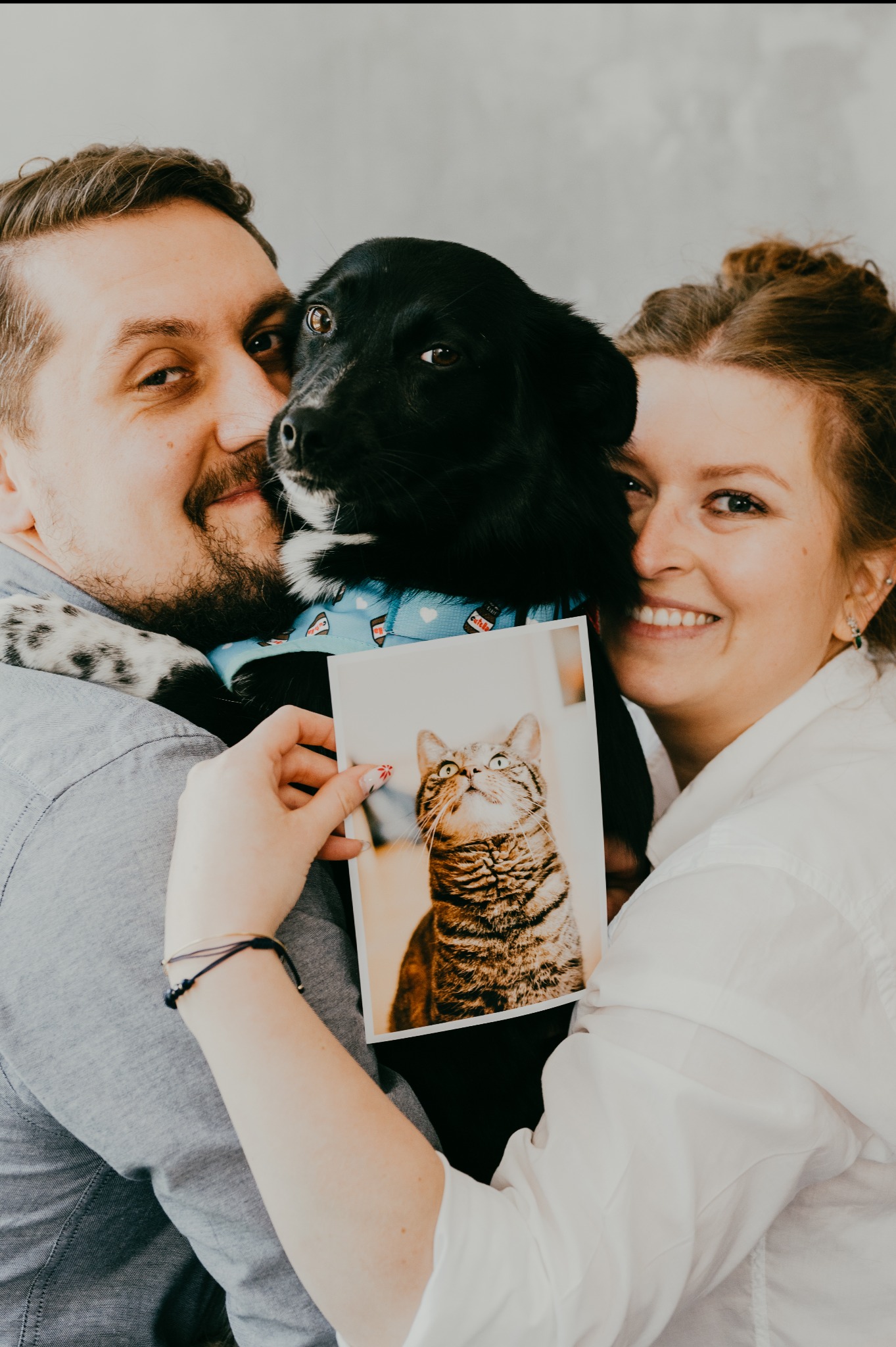 Uśmiechnięta para obejmuje czarnego psa z niebieską apaszką, trzymając zdjęcie kota w ramce.