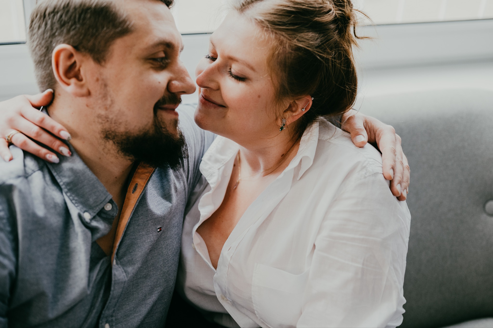 Uśmiechnięta para, on z brodą w szarej koszuli, ona w białej, obejmująca się czule, zbliżająca się do pocałunku, z kolczykiem typu helix w uchu, na neutralnym tle.