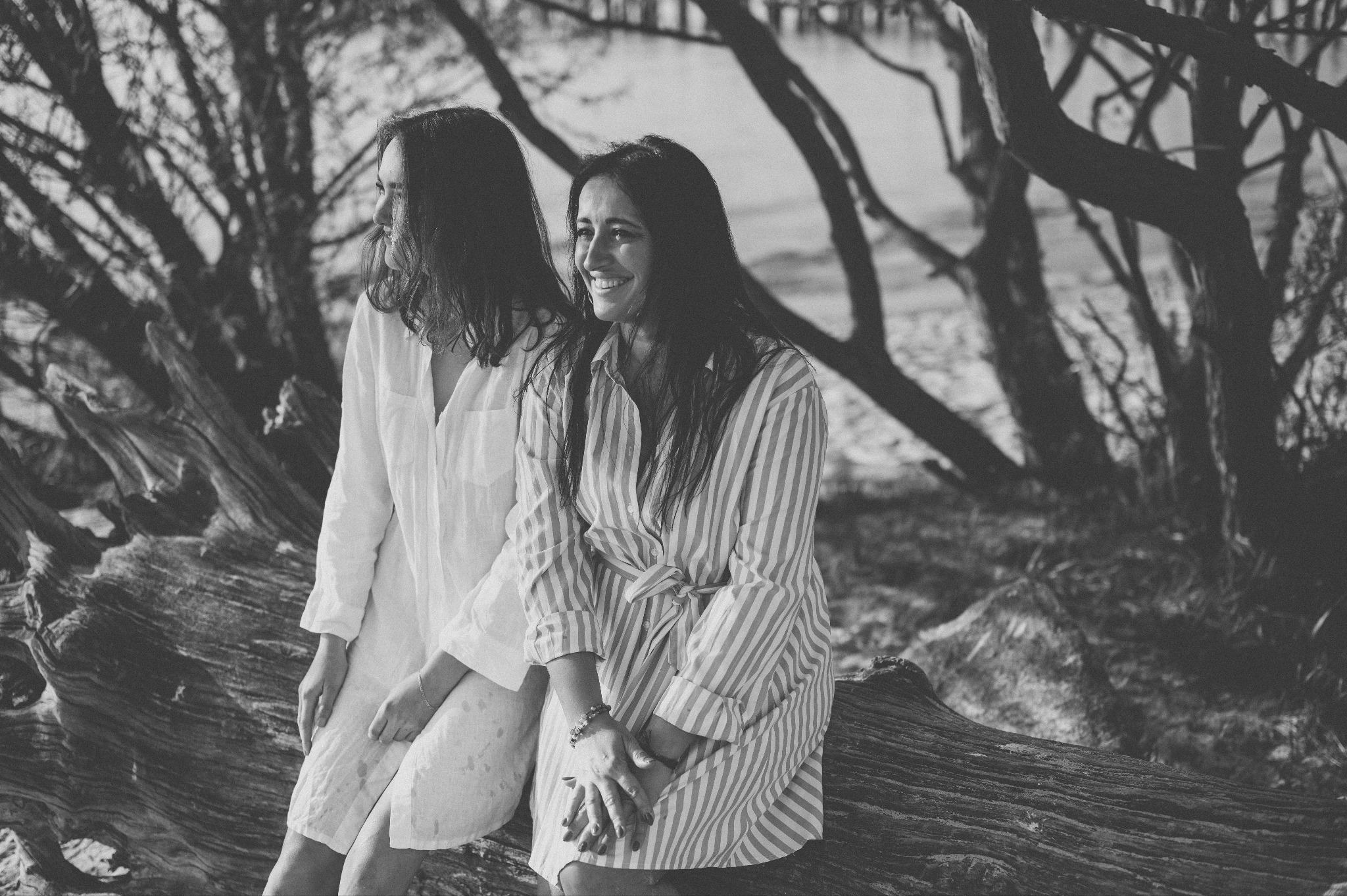 Dwie uśmiechnięte kobiety siedzą na spróchniałym pniu drzewa na tle plaży i drzew, ubrane w zwiewne letnie sukienki w biało-szarej tonacji, uchwycone w naturalnym świetle.