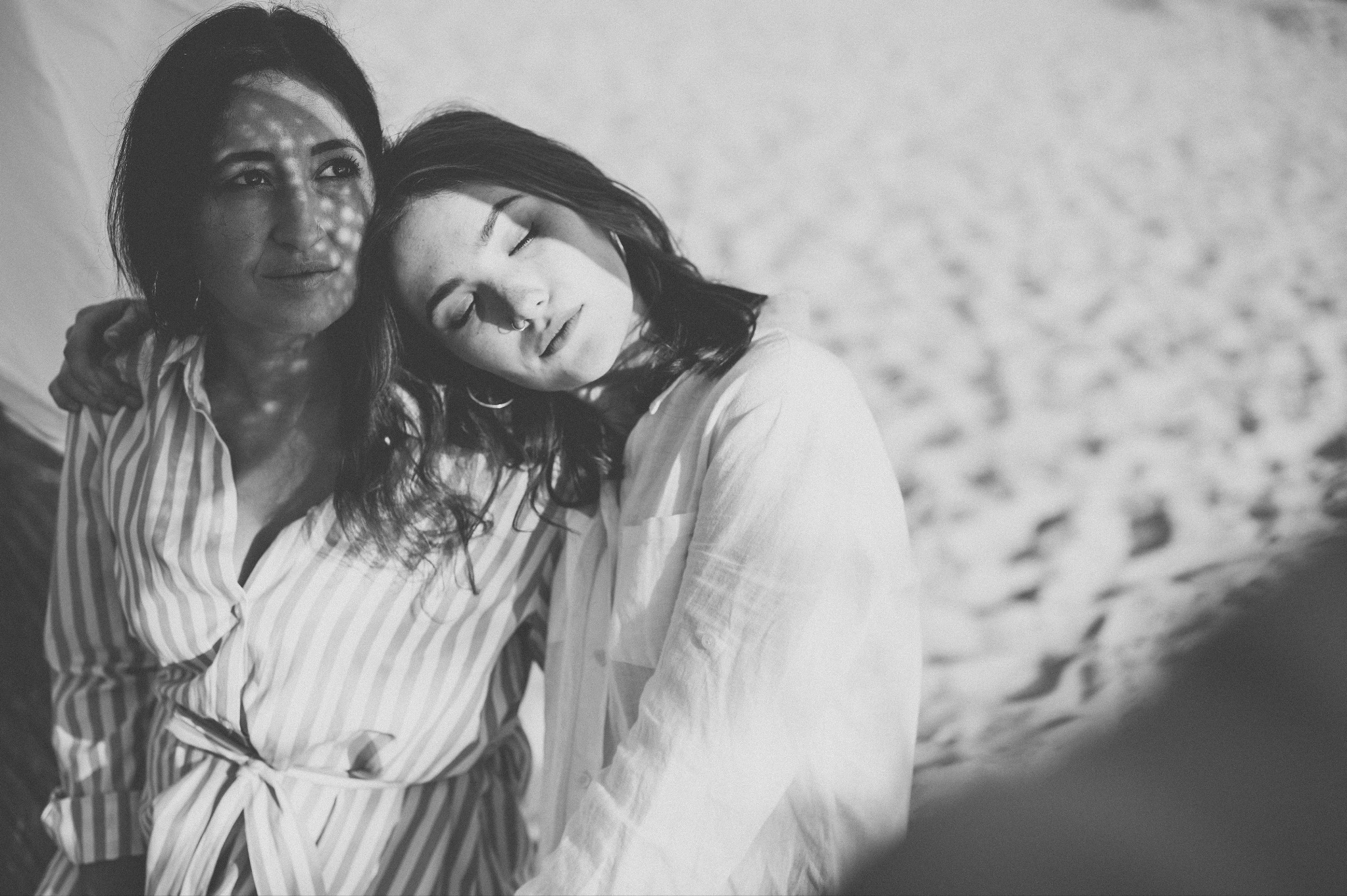 Czarno-białe zdjęcie dwóch kobiet w bliskim uścisku na tle jasnego, rozproszonego światła. Jedna kobieta opiera głowę na ramieniu drugiej, ma zamknięte oczy i kolczyk w nosie. Druga kobieta patrzy...