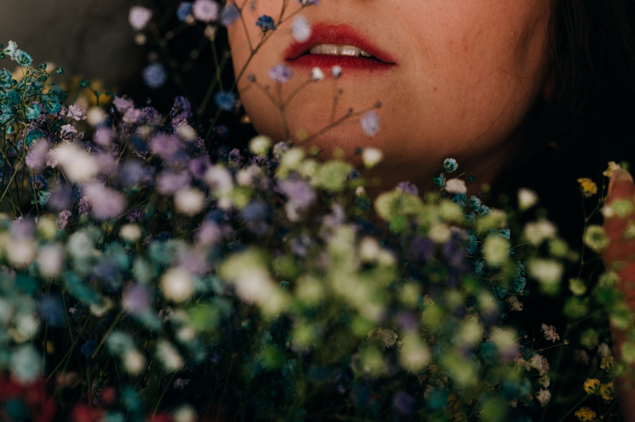 Fragment twarzy kobiety z czerwonymi ustami, otoczony bukietem wielobarwnego gipsówki w odcieniach fioletu, zieleni i błękitu, tworzący intymny portret.