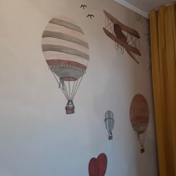 BARBARA KULESZA DOM I WNĘTRZE - Ściana w pokoju dziecięcym z ręcznie malowanymi motywami: balony na ogrzane powietrze, samolot dwupłatowy i ptaki w locie, całość utrzymana w stonowanej kolorystyce, widoczne żółte zasłony w tle.