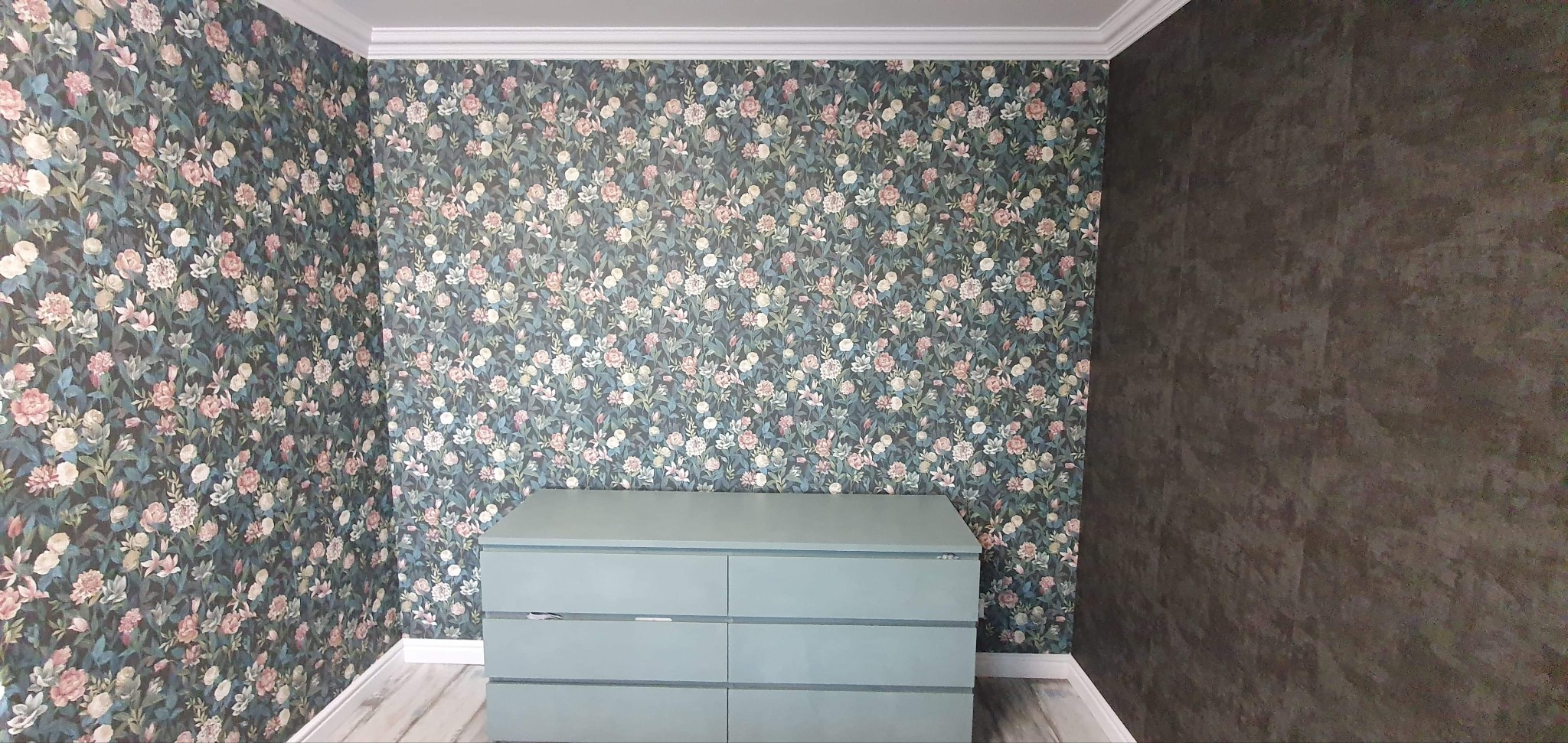 Pokój z tapetą w drobne kwiaty i komodą w kolorze pastelowej zieleni, jedna ściana pomalowana na ciemny, grafitowy kolor z efektem struktury.
