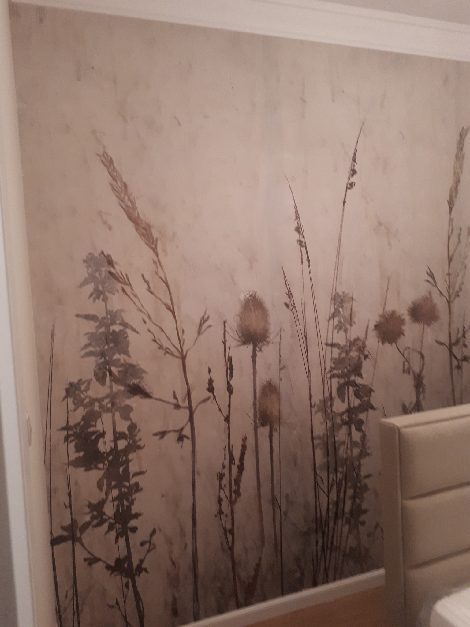 Fototapeta w sypialni przedstawiająca motyw polnych traw i ostów na beżowym tle z delikatną teksturą, widoczny fragment zagłówka łóżka.