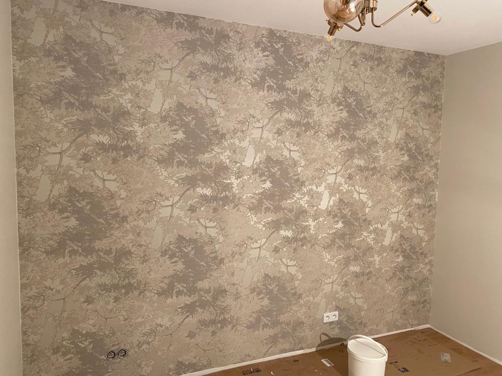 Ściana w sypialni pokryta tapetą z motywem drzew w odcieniach szarości i beżu, widoczna lampa sufitowa i fragment podłogi zabezpieczonej kartonem.