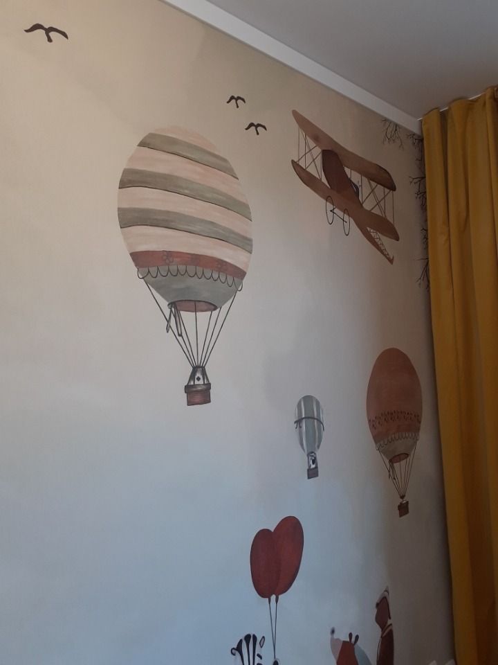 Ściana w pokoju dziecięcym z ręcznie malowanymi motywami: balony na ogrzane powietrze, samolot dwupłatowy i ptaki w locie, całość utrzymana w stonowanej kolorystyce, widoczne żółte zasłony w tle.
