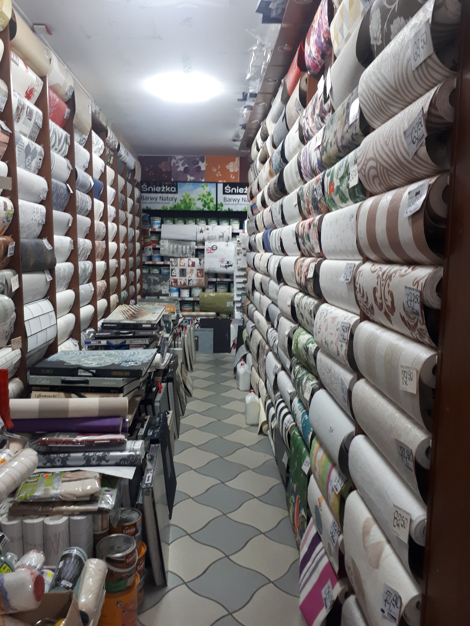 Wnętrze sklepu z tapetami: długi korytarz pomiędzy regałami wypełnionymi rolkami tapet o różnych wzorach i kolorach, w tle widoczne farby i inne akcesoria malarskie, podłoga w szaro-białą kratę.
