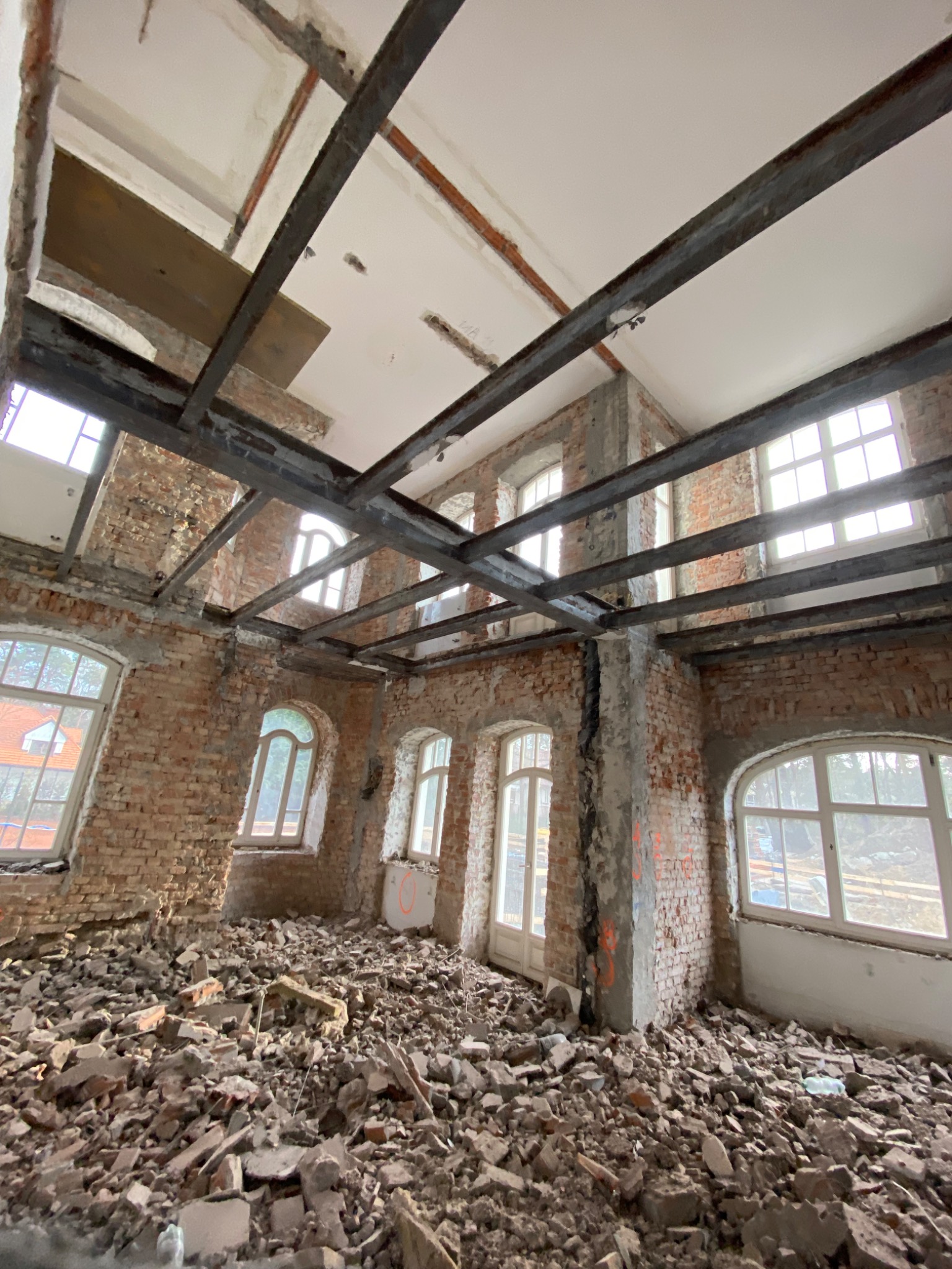 Wnętrze budynku w trakcie rozbiórki, widoczne ceglane ściany z oknami, metalowa konstrukcja stropu i gruz na podłodze.