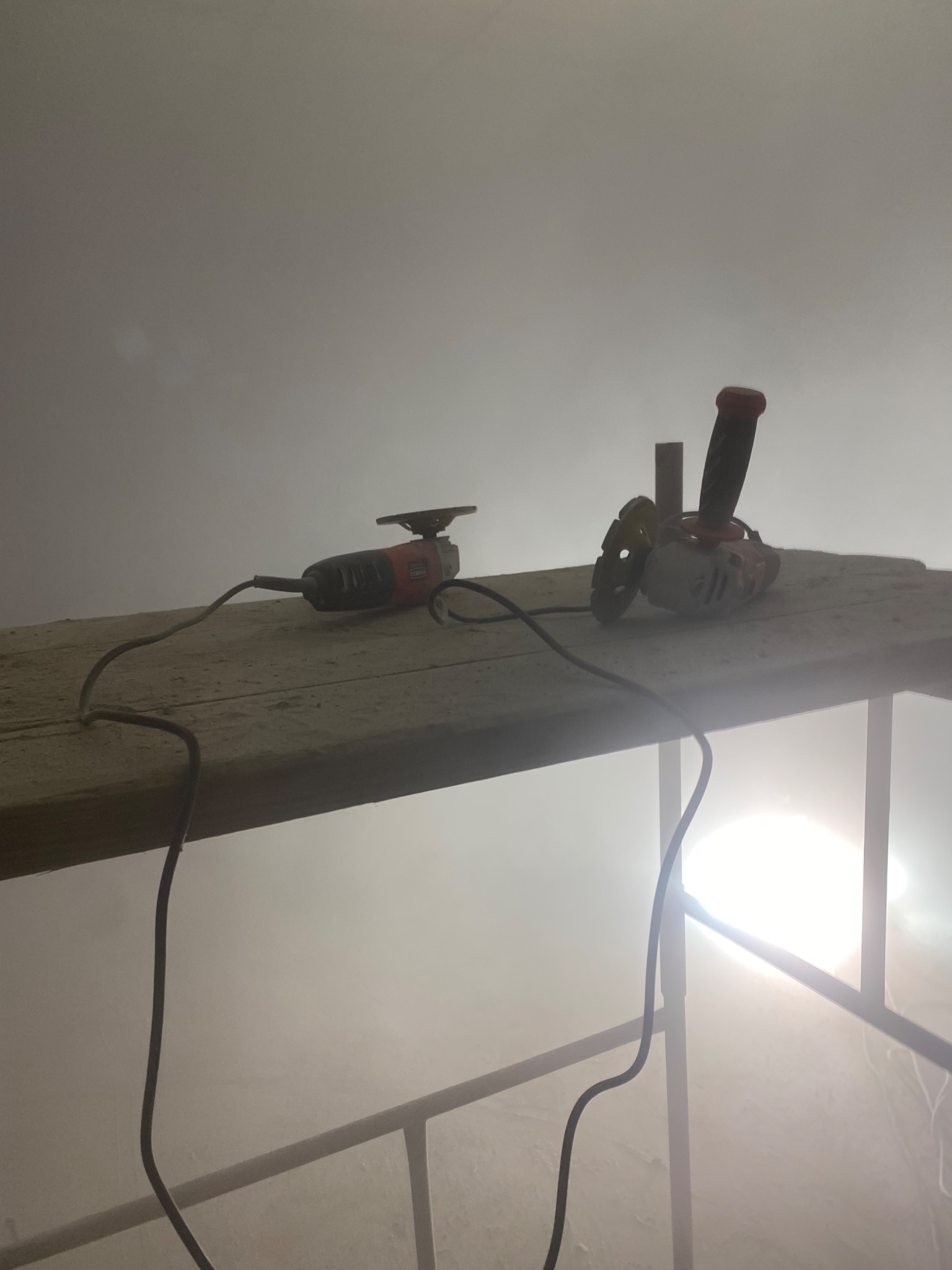 Dwie szlifierki kątowe, jedna z tarczą diamentową, stoją na zakurzonej desce rusztowania w zapylonym pomieszczeniu.