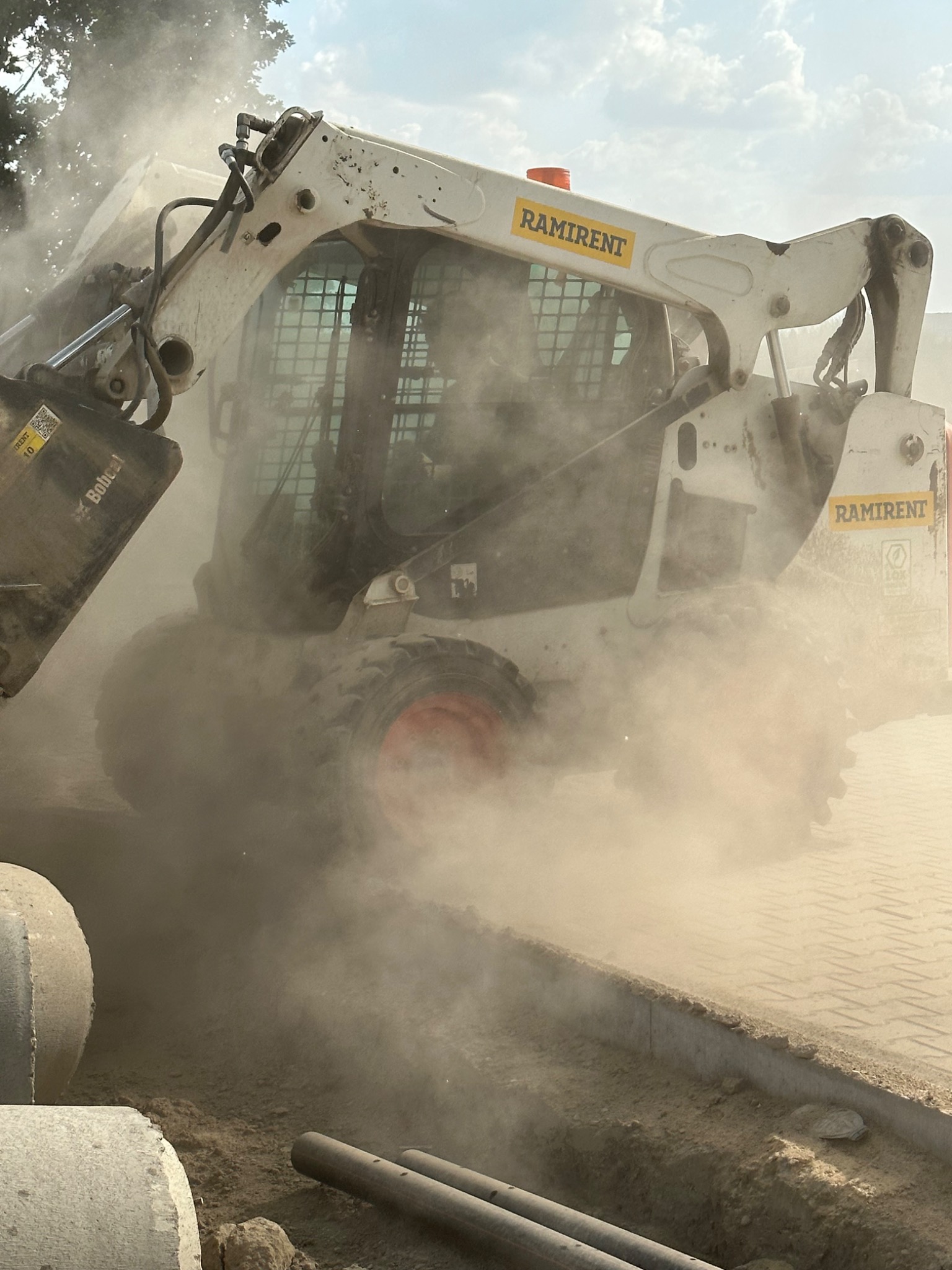 Bobcat z łyżką w chmurze pyłu, widoczny napis Ramirent, obok leżą betonowe kręgi