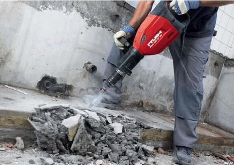 Pracownik w szarych spodniach używa czerwonego młota pneumatycznego Hilti do skuwania betonowej ściany, widoczne rury instalacyjne w ścianie i gruz.