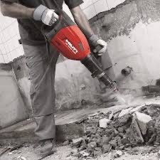 Mężczyzna w szarych spodniach i rękawicach ochronnych używa czerwonego młota pneumatycznego Hilti do kruszenia ściany pokrytej resztkami płytek, widoczne gruz i kurz.