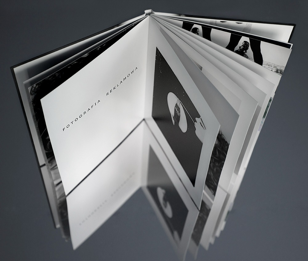 Otwarty album z czarno-białymi zdjęciami, widoczne strony z napisami 'Fotografia Reklamowa' i 'Kandydacie Fotograficzny', ujęcie z góry, szare tło.